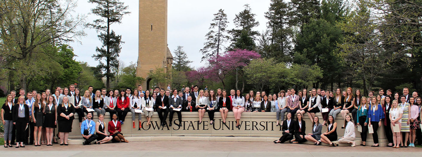 Graduating honors students sitting on and round the Iowa State University sign in front of the Campanile.